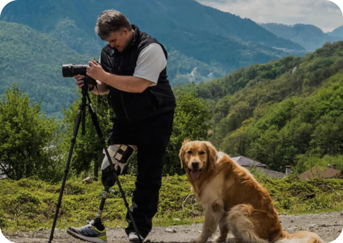 мужчина с протезом делает фотографии на фоне гор рядом с собакой